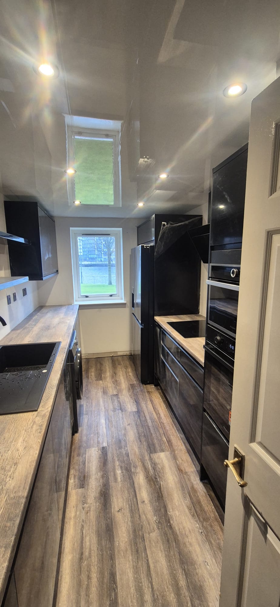 Modern galley kitchen with black gloss units, wood-effect worktop, LVT flooring and integrated appliances